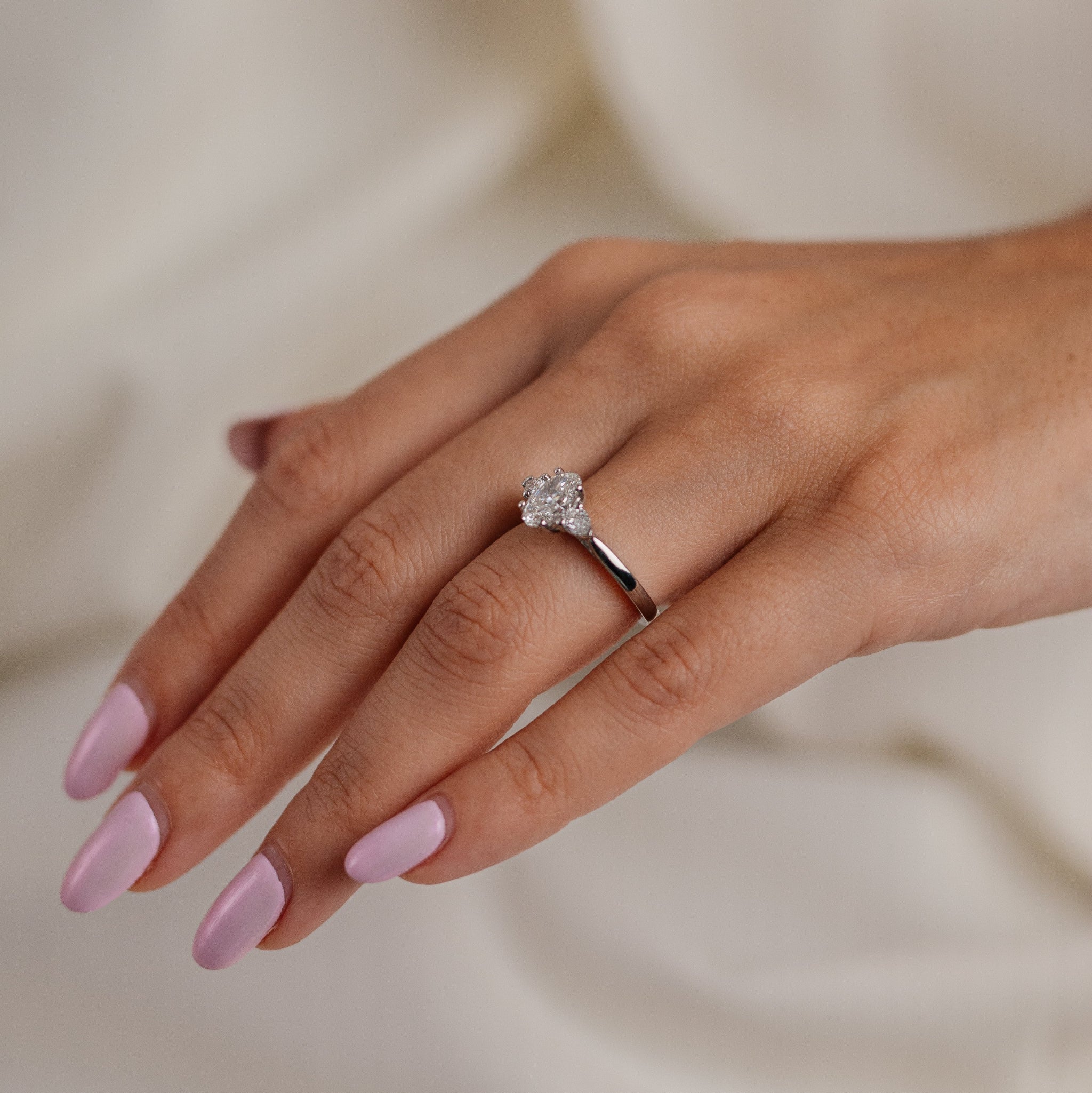 Hand wearing a diamond ring with pink nail polish on a neutral background