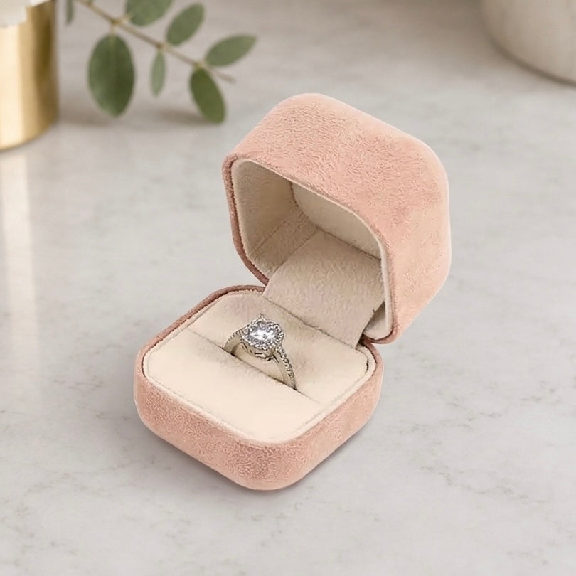 A silver ring with a clear center stone is displayed inside an open blush pink velvet ring box on a light marble surface.