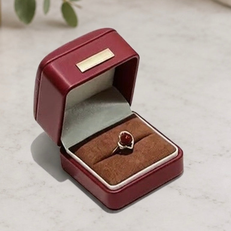 A small open burgundy ring box with a velvet-lined cushion holding a ring with a round red stone and a silver-toned setting, photographed on a light marble-like surface.