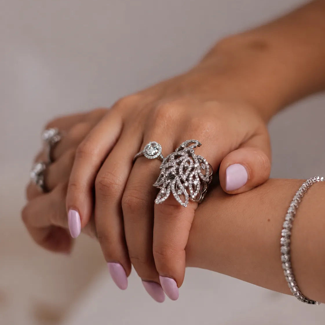 A close-up of a person's hands and wrists showcasing jewellery, featuring a large leaf-shaped ring with pavé stones, a delicate solitaire ring, a matching pavé bracelet, and a small ring on a gold band.