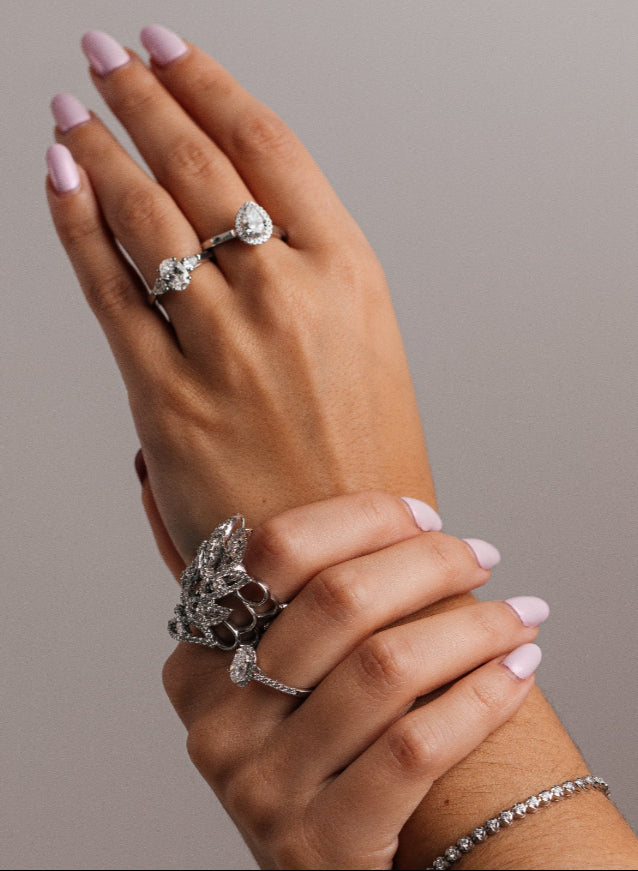 Hand model wearing multiple diamond platinum rings with pink nail polish on a neutral background