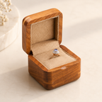 A gold ring with a round blue gemstone and small clear stones, displayed inside an open wooden ring box with a beige velvet interior on a light beige surface.