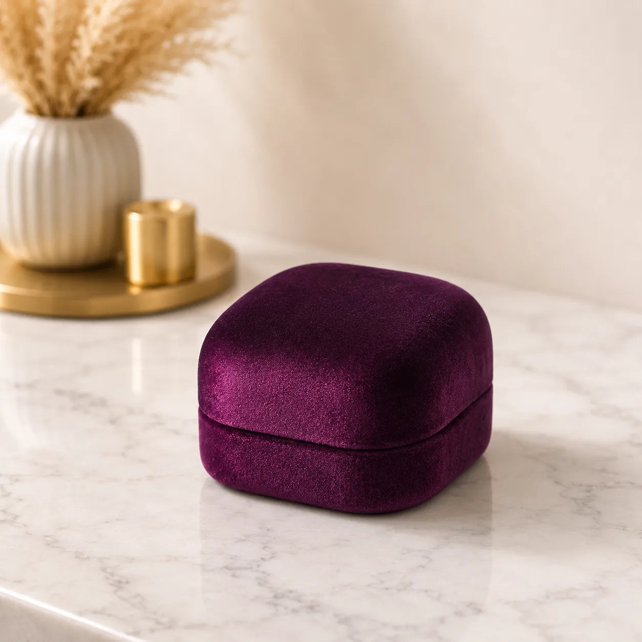 A small square velvet jewelry box in deep purple sits on a white marble surface, with a cream vase of dried grass and a gold candle holder blurred in the background.