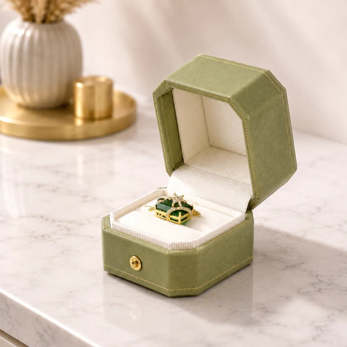 An open sage green velvet ring box with a white velvet interior, holding a gold ring with a green and white patterned center, set on a white marble surface with a blurred decorative background.