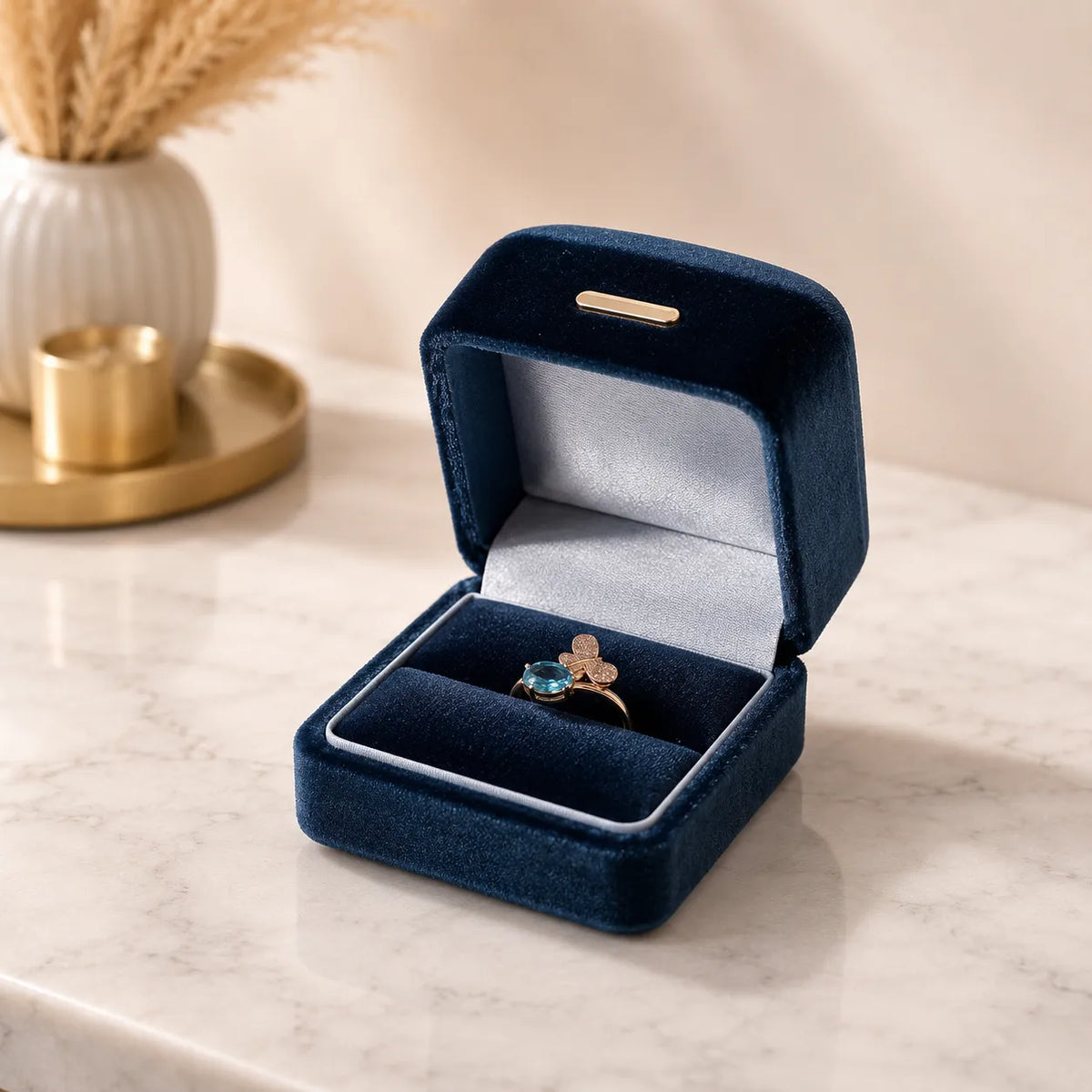 A gold ring with a blue gemstone and a clover motif sits inside an open dark blue velvet ring box on a light marble surface.