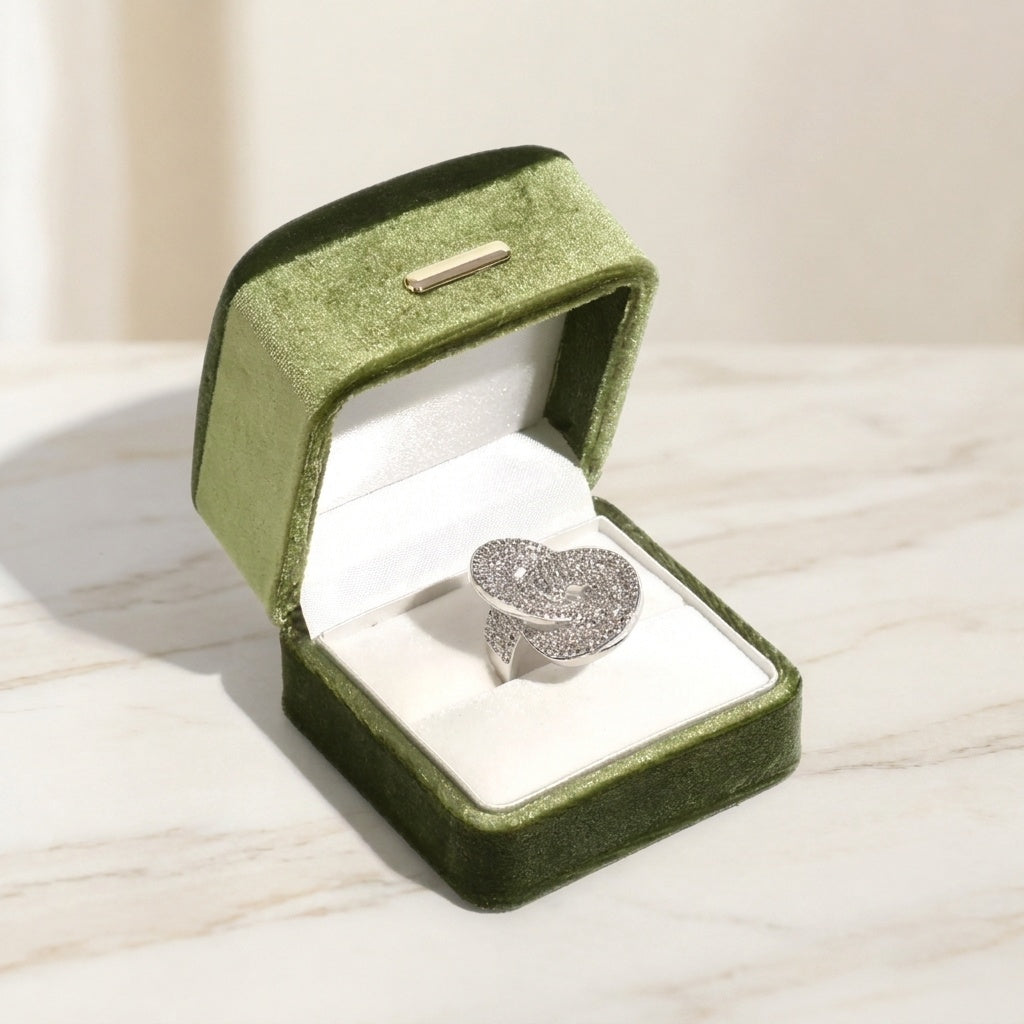 A silver ring with a textured, diamond-like pavé design rests inside an open olive green velvet ring box on a light marble surface.