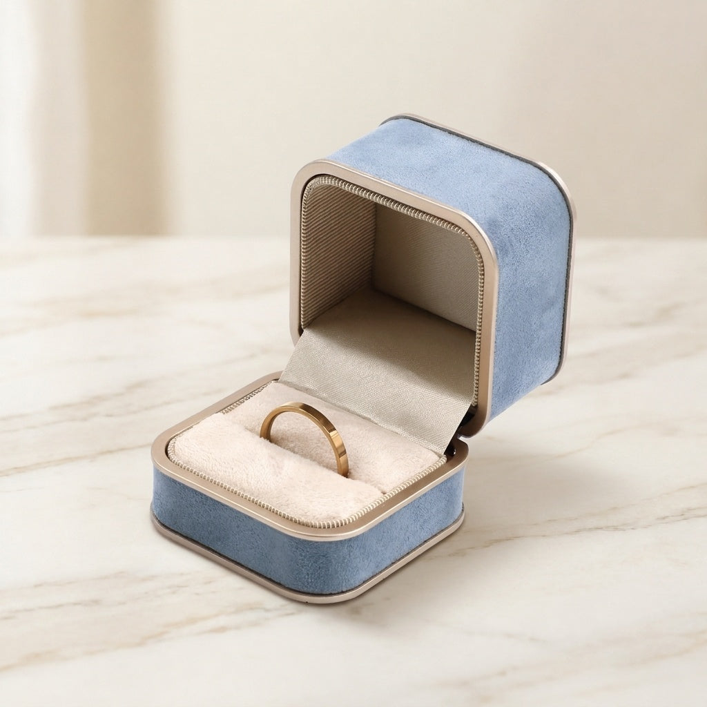 A small open jewelry box with a light blue velvet exterior, beige interior lining, and a gold ring displayed inside, set on a light marble surface against a soft cream background.