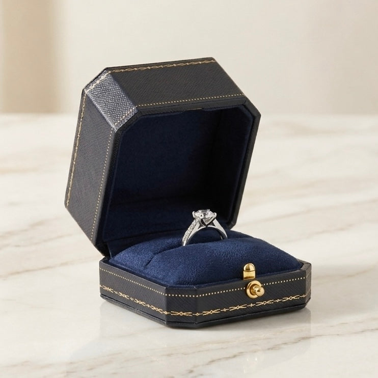 A silver ring with a clear gemstone displayed inside an open black jewelry box with a navy blue velvet lining, set on a light marble surface.