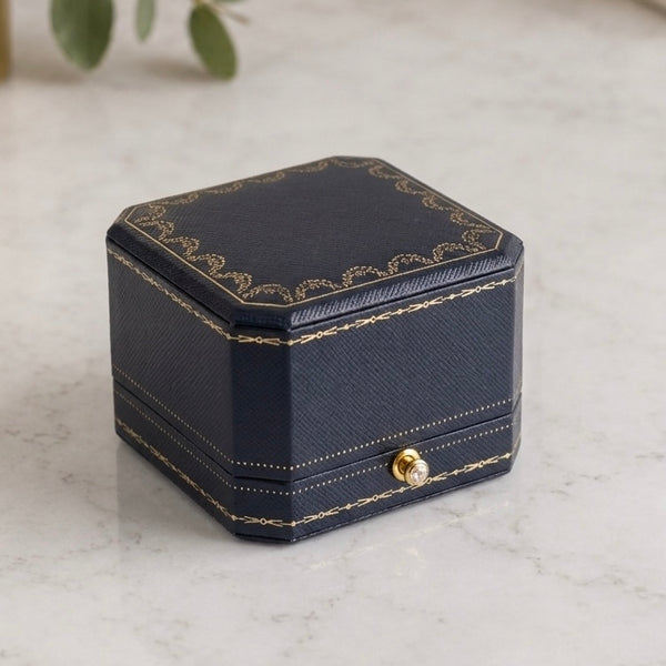A dark navy blue textured jewelry box with gold decorative trim and a small gold clasp, set on a light marble surface.